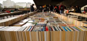 Choose books. Three book stands with books standing in a square shape surrounding many more books that are laying down, a person is looking at the books, another is reading a book on a chair and a third is speaking with him.