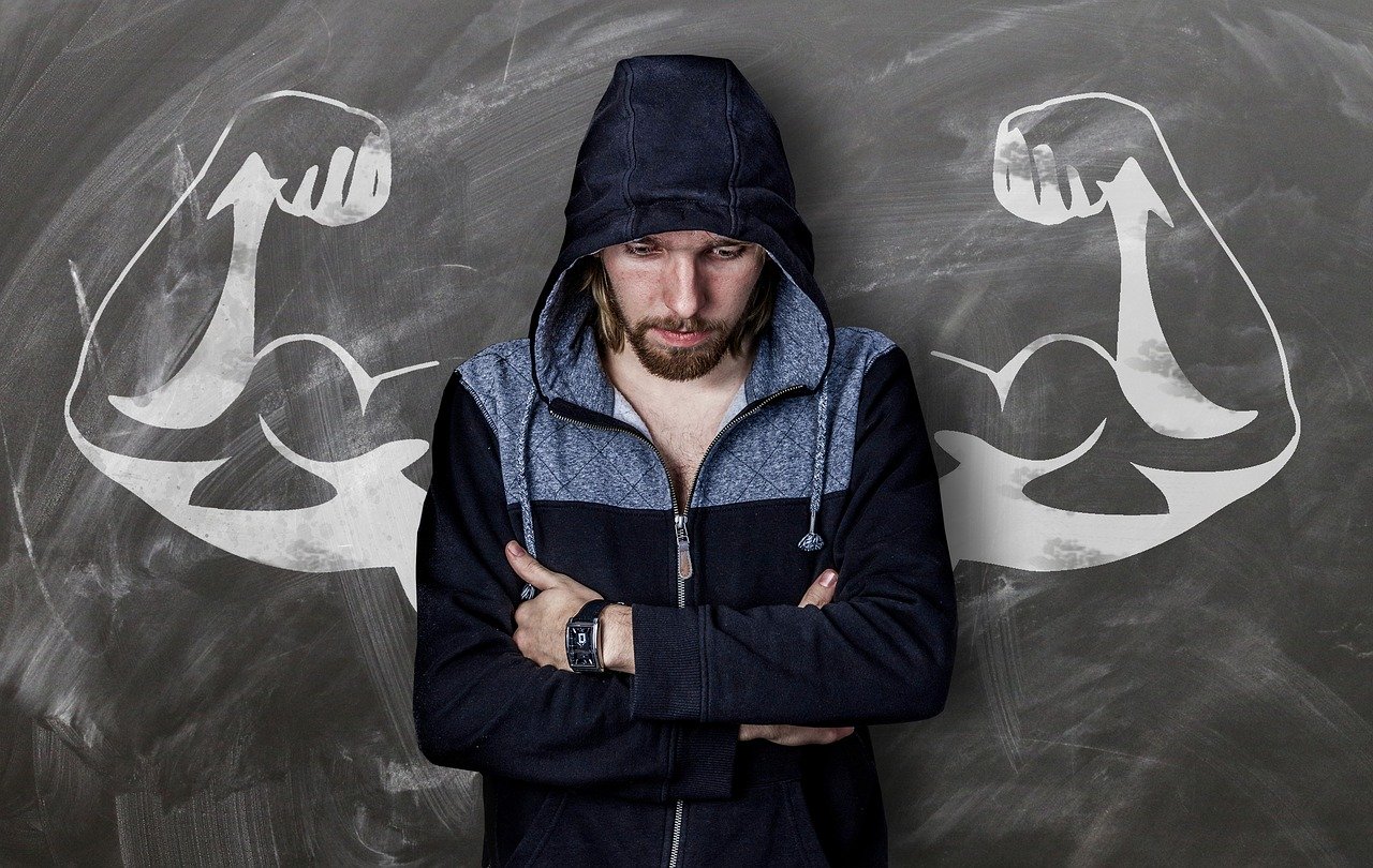 Kelly Clarkson - Stronger. A man with a part dark part light blue hoodie is standing with his arms crossed looking down, at the background is a whiteboard at which a muscular extension of his arms appears as drawed